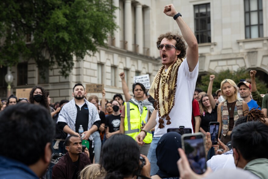 Ammer Qaddumi is wearing a white t-shirt and a brown and beige keffiyeh while speaking at a protest on the UT Austin campus. He has one fist raised in the air. 