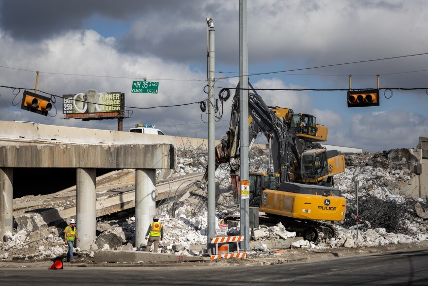 Excavators demolishing the northbound I-35 bridge over Howard Lane. Construction workers in high-visibility gear are standing by. Traffic lights hang above, with visible signage for I-35 and a billboard in the background that has a picture of a wooly mammoth. 