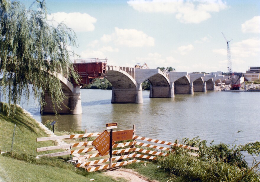 The partially constructed Congress Avenue Bridge over what was then known as Town Lake. A barricade with orange and white striped signs indicates the bridge is closed to pedestrian traffic. A crane is visible on the right side, with buildings in the background. 