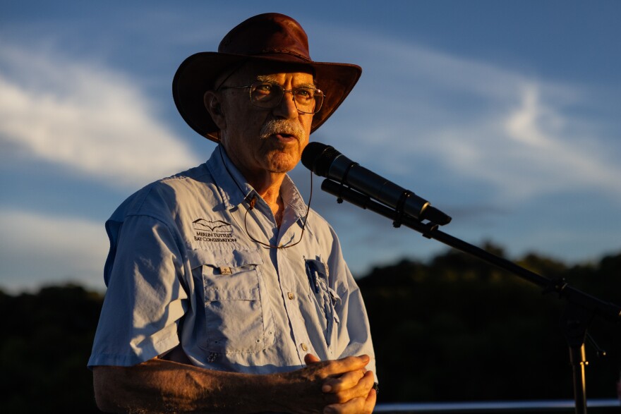 Merlin Tuttle, a bat ecologist and conservationist, wears a wide-brimmed hat and a light blue shirt with "Merlin Tuttle's Bat Conservation" embroidered on as he speaks into a microphone outdoors.