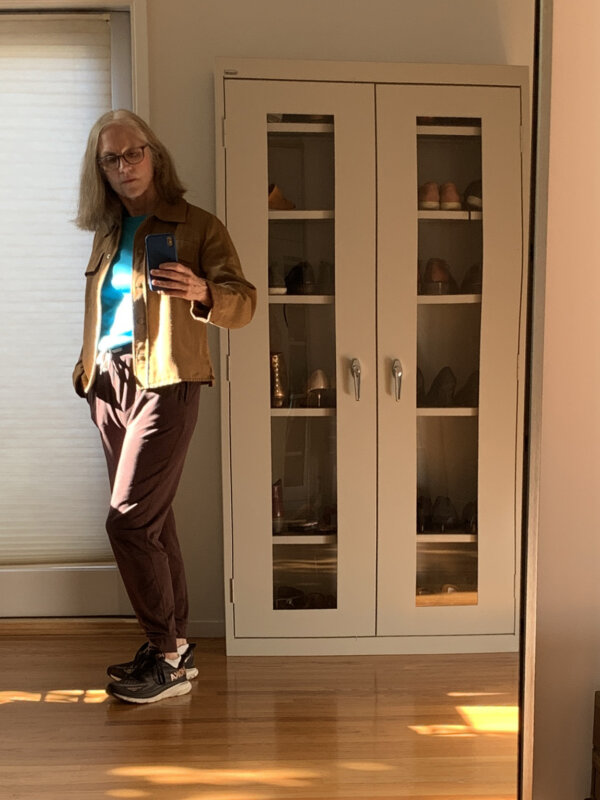 Older woman standing in afternoon light wearing an ochre jacket and a turquoise t-shirt