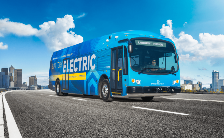 A blue Proterra ZX5 bus. The bus is seen driving down the road wrapped in a sign that says, "Battery electric." A city skyline is in the background. 