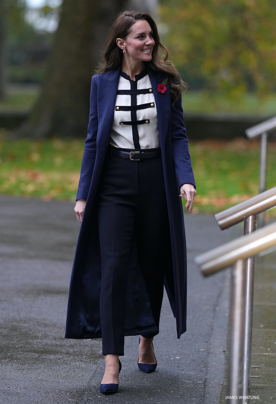 Kate Middleton wearing a navy coat over a white blouse with navy accents, paired with tailored navy trousers and heels, walking outdoors and smiling.