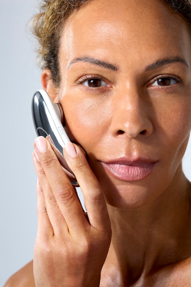 A woman holds a small device up to her cheek. One side is smooth and chrome, and the side touching her face has two metal nodes that conduct microcurrent electricity in and out of the skin.