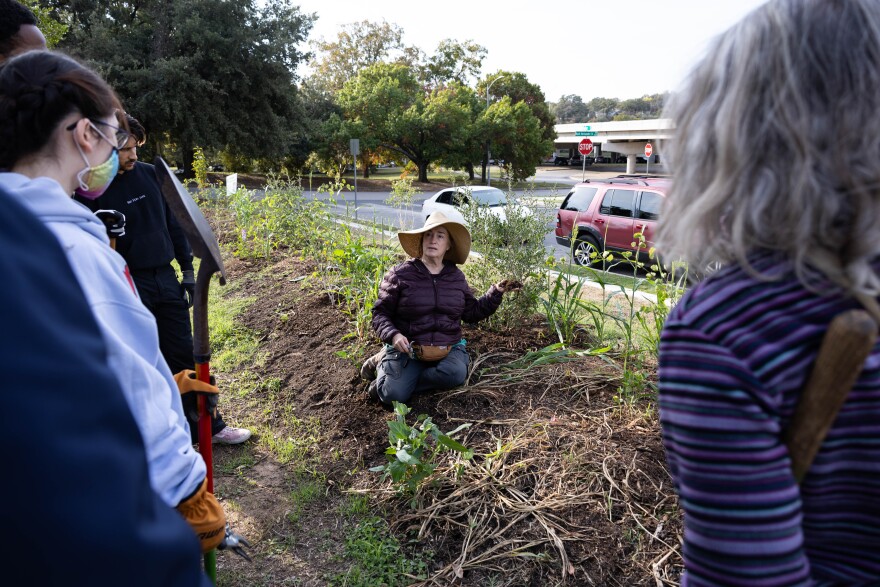 A woman kneels down in a grassy area as she speak to people standing around her.