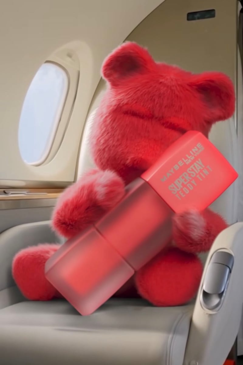 A pink, furry teddy bear is sitting on a plane with the window open and cradling an oversized tube of Maybelline Superstay Teddy Tint lip oil.