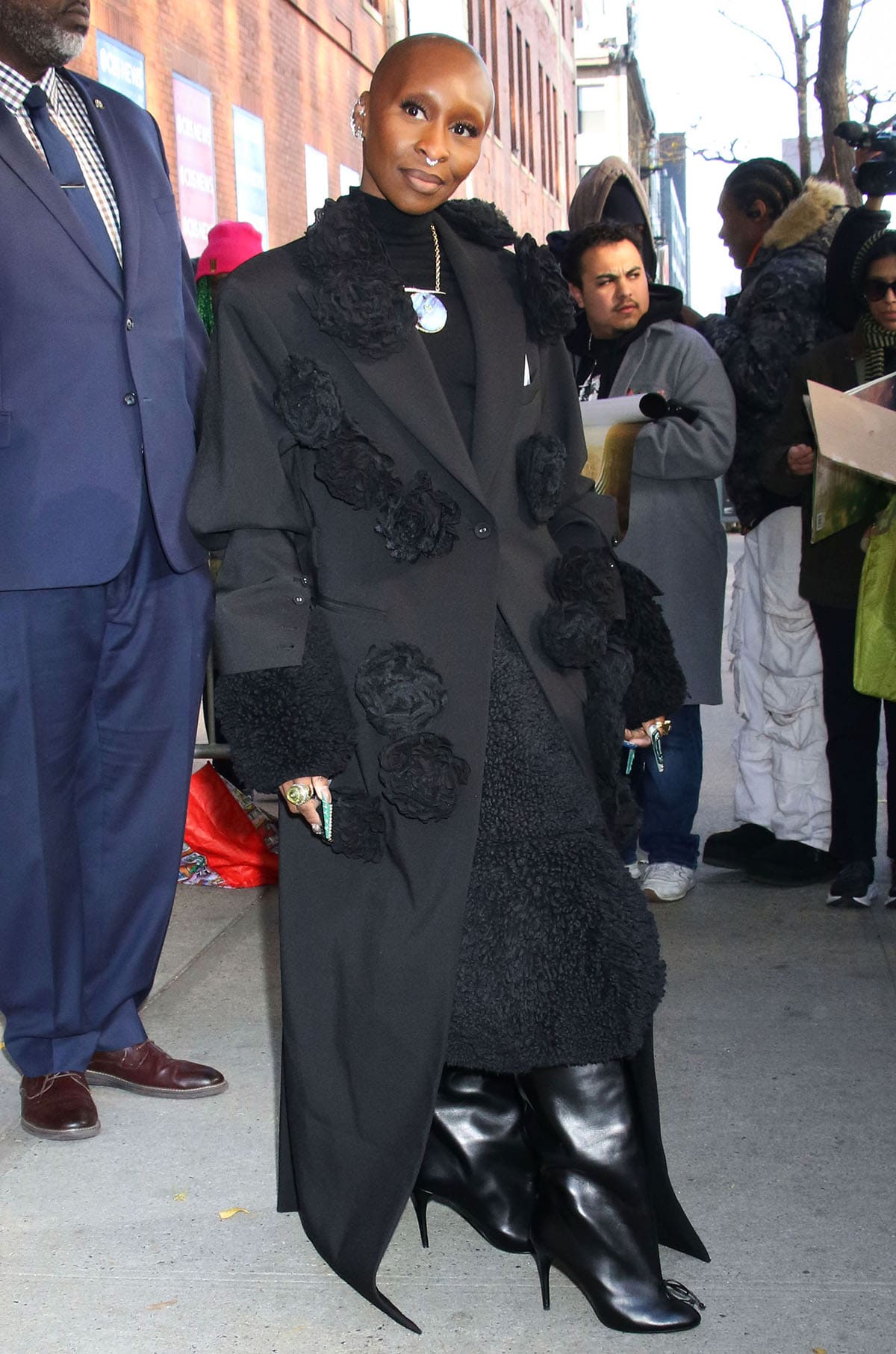 Cynthia Erivo wears a black coat with 3D flower accents by Rokh over a black dress with a see-through bodice and fur trims