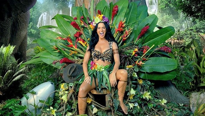 A glamazon Katy Perry sitting on a throne of palm leaves and tropical flowers in the official music video for "Roar"